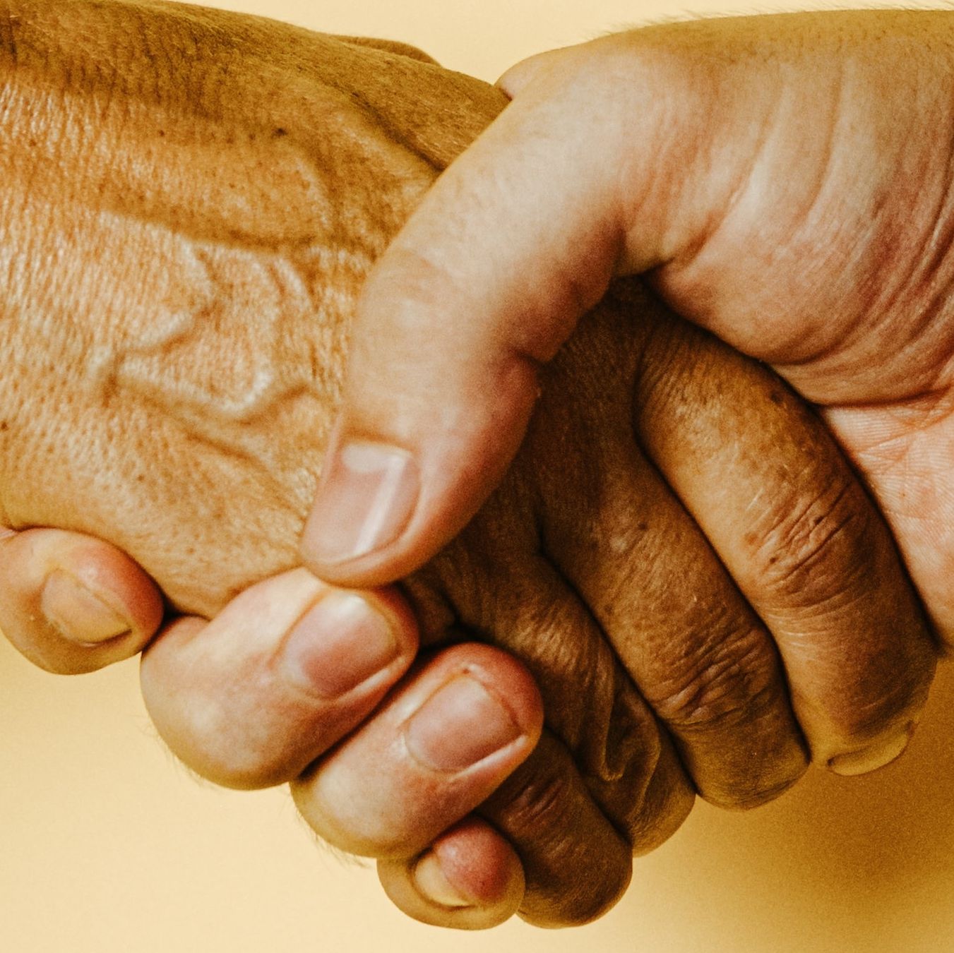 Eine Nahaufnahme von zwei Händen, eine gealtert und eine jünger, die sich in einer Geste der Unterstützung und Fürsorge halten.