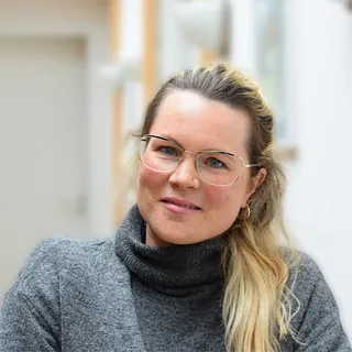 Frau mit blonden Haaren, Brille und grauem Rollkragenpullover.
