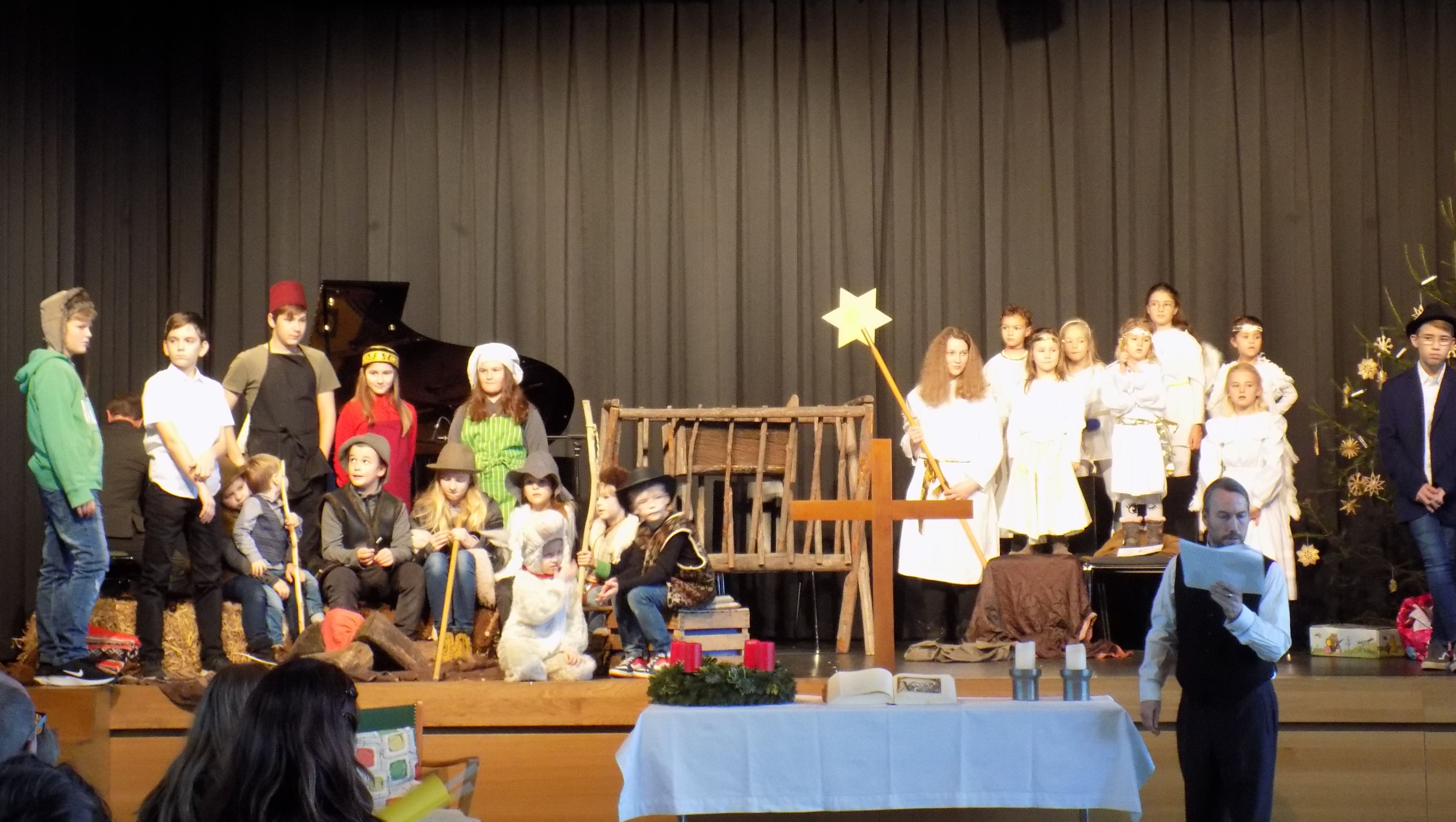  Kinder führen ein Krippenspiel auf einer Bühne mit Weihnachtsdekoration auf.