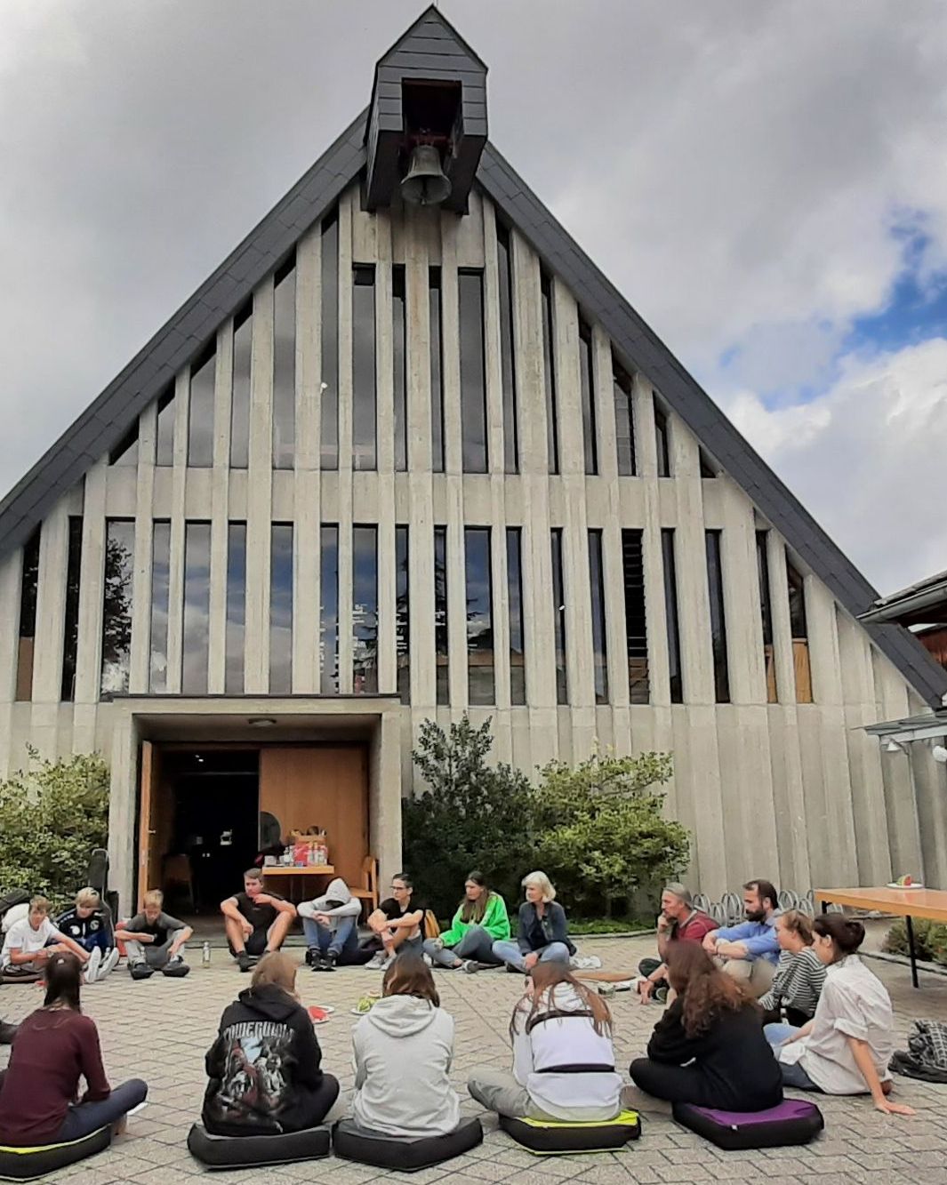 Gruppe sitzt im Kreis vor moderner Kirche mit Holzakzenten.