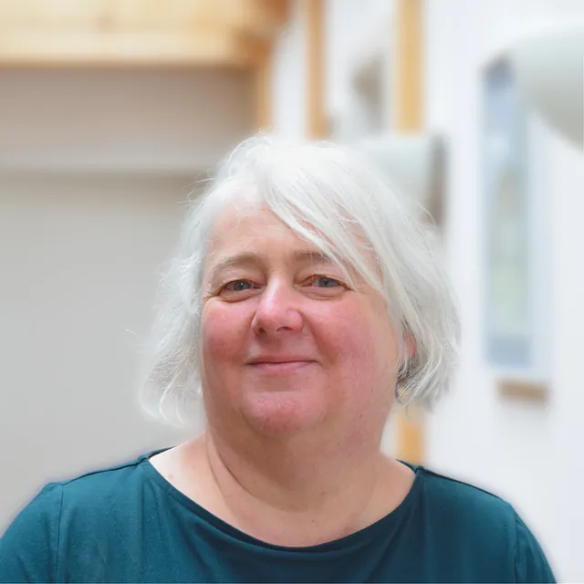 Frau mit kurzen weißen Haaren, die ein petrolfarbenes Shirt trägt und leicht lächelt.